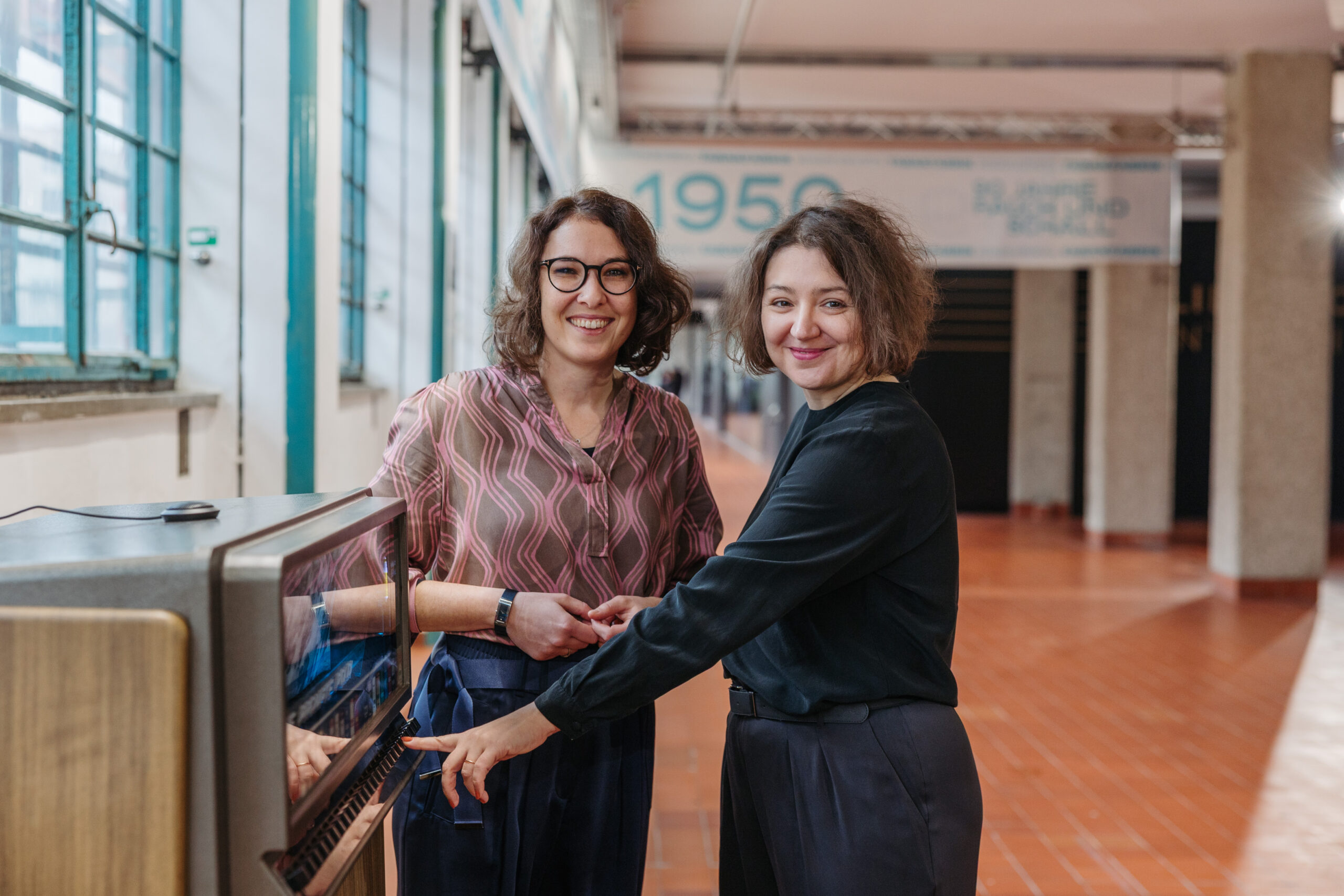 Zwei Personen stehen bei der KI-gesteuerten Poldi, dem historischen Zigarettenautomaten, der auf Knopfdruck sein Wissen über 90 Jahre Tabakfabrik Linz erzählt. Er ist Teil der Ausstellung "90 Jahre Rauch & Schall".