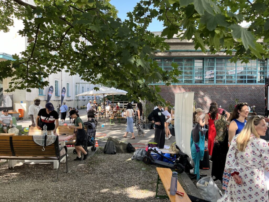 Einige Menschen halten sich im Innenhof der Tabakfabrik auf, das Wetter ist gut, der Baum bietet Schatten, Tische sind gedeckt, es wird geplaudert, getrunken...