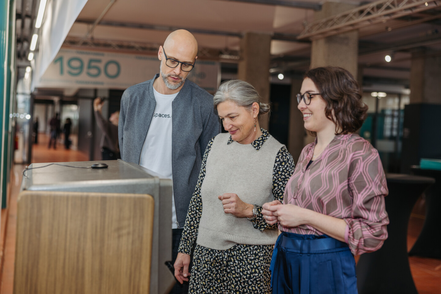 Drei Personen stehen bei der KI-gesteuerten Poldi, dem historischen Zigarettenautomaten, der auf Knopfdruck sein Wissen über 90 Jahre Tabakfabrik Linz erzählt. Er ist Teil der Ausstellung "90 Jahre Rauch & Schall".
