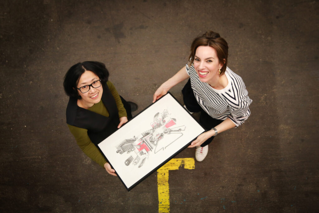 Künstlerin Xian Zheng und Maximiliane Buchner von der Kunstrampe blicken nach oben in die Kamera und halten gemeinsam ein Kunstwerk.